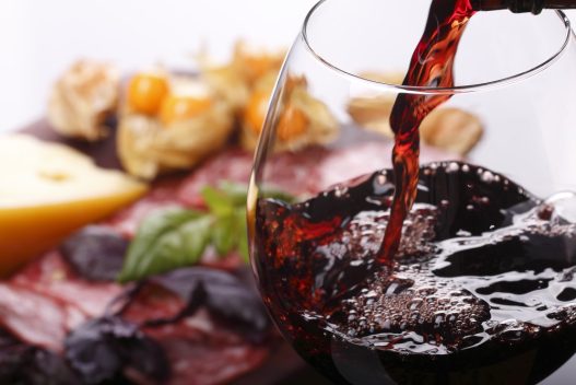 Der hældes vin i et vinglas med mad i baggrunden
